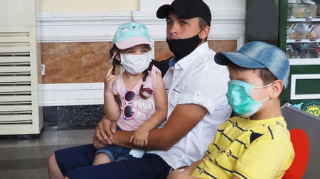 Father with daughter in his arms, son sits wearing masks at station or at the airport, waiting for the train. Family in face mask. Virus. Coronavirus and flu pandemic. Safe travel with child and babyの写真素材