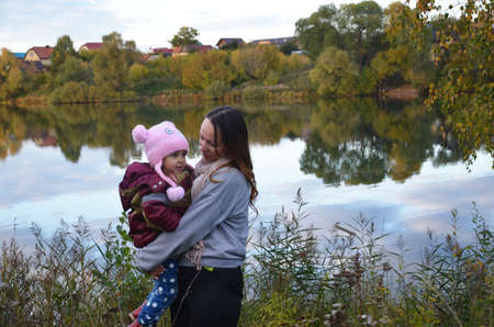 mom and daughter walk by the lake in autumn. Against a background of autumn colours and leaves, a mother smiling next to her daughter. The trees are reflected in the lake.の写真素材
