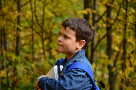 A boy after school hours walks in the autumn park in a denim jacket, with textbooks and notepads, notebooks in his hands in the park among the trees. The concept of heavy learning, a lot of homeworkの写真素材
