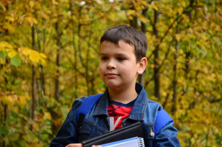 A boy after school hours walks in the autumn park in a denim jacket, with textbooks and notepads, notebooks in his hands in the park among the trees. The concept of heavy learning, a lot of homeworkの写真素材