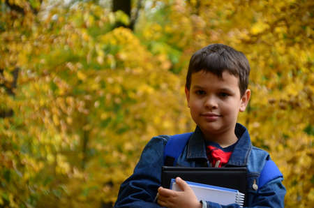 A boy after school hours walks in the autumn park in a denim jacket, with textbooks and notepads, notebooks in his hands in the park among the trees. The concept of heavy learning, a lot of homeworkの写真素材