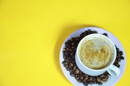 warm cup of coffee cappuccino with cream in a white cup on an yellow background with coffee beans with a teaspoon. top viewの写真素材