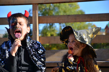 children in halloween costumes on holiday a girl in a witch suit a black hat on the head with black Halloween make-up, boy in devil halloween costume with horns zombie eating sweets jelly wormsの写真素材