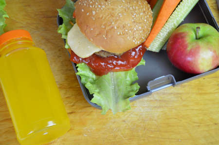 Homemade Hamburgers with lettuce in lunchbox on the table. with bright yellow orange juice. with banana nuts. Sliced vegetables carrots and cucumber. Opposite the school board. Snack. School lunchの写真素材