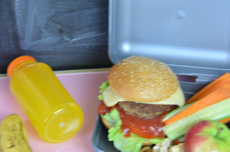 Homemade Hamburgers with lettuce in lunchbox on the table. with bright yellow orange juice. with banana nuts. Sliced vegetables carrots and cucumber. Opposite the school board. Snack. School lunchの写真素材