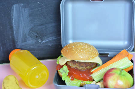 Homemade Hamburgers with lettuce in lunchbox on the table. with bright yellow orange juice. with banana nuts. Sliced vegetables carrots and cucumber. Opposite the school board. Snack. School lunchの写真素材