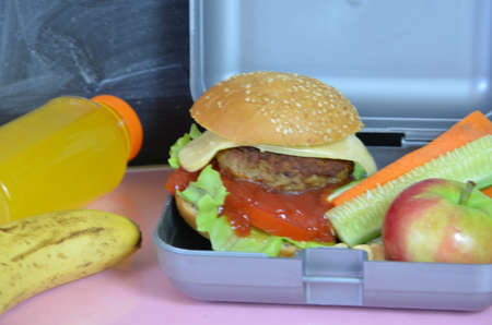 Homemade Hamburgers with lettuce in lunchbox on the table. with bright yellow orange juice. with banana nuts. Sliced vegetables carrots and cucumber. Opposite the school board. Snack. School lunchの写真素材