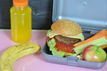 Homemade Hamburgers with lettuce in lunchbox on the table. with bright yellow orange juice. with banana nuts. Sliced vegetables carrots and cucumber. Opposite the school board. Snack. School lunchの写真素材
