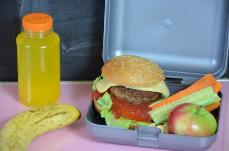 Homemade Hamburgers with lettuce in lunchbox on the table. with bright yellow orange juice. with banana nuts. Sliced vegetables carrots and cucumber. Opposite the school board. Snack. School lunchの写真素材