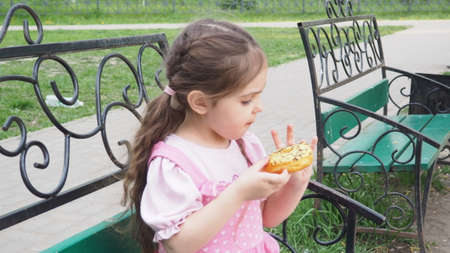 girl on a park bench eating a donut with yellow frosting. happy childhood, outdoor recreation, walks. Cheerful Child Sitting on Park Benchの写真素材