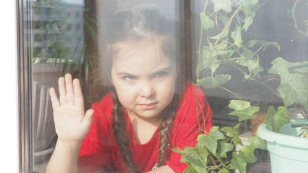 Beautiful little girl smiling and watching out the window. A child looks out the window. Portrait of cheerful kid. self distancing at home during Corona virus. Quarantine Covid-19 World Pandemic.の写真素材