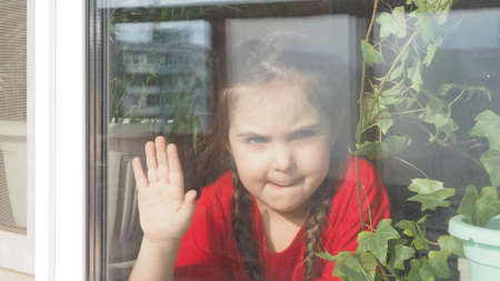 Beautiful little girl smiling and watching out the window. A child looks out the window. Portrait of cheerful kid. self distancing at home during Corona virus. Quarantine Covid-19 World Pandemic.の写真素材