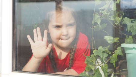 Beautiful little girl smiling and watching out the window. A child looks out the window. Portrait of cheerful kid. self distancing at home during Corona virus. Quarantine Covid-19 World Pandemic.の写真素材