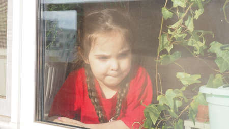 Beautiful little girl smiling and watching out the window. A child looks out the window. Portrait of cheerful kid. self distancing at home during Corona virus. Quarantine Covid-19 World Pandemic.の写真素材