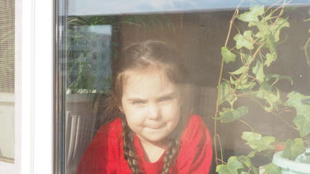 Beautiful little girl smiling and watching out the window. A child looks out the window. Portrait of cheerful kid. self distancing at home during Corona virus. Quarantine Covid-19 World Pandemic.の写真素材