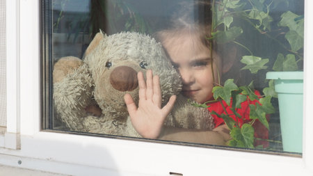 Beautiful little girl smiling and watching out the window. A child looks out the window. Portrait of cheerful kid. self distancing at home during Corona virus. Quarantine Covid-19 World Pandemic.の写真素材
