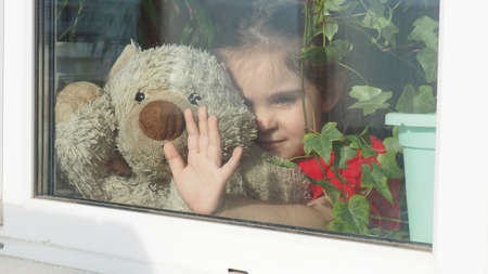 Beautiful little girl smiling and watching out the window. A child looks out the window. Portrait of cheerful kid. self distancing at home during Corona virus. Quarantine Covid-19 World Pandemic.の写真素材