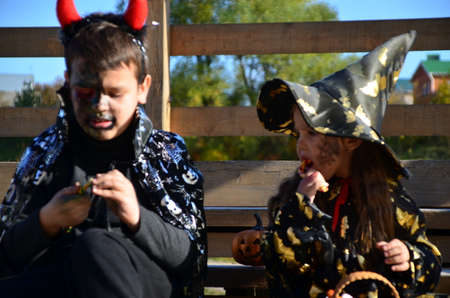children in halloween costumes on holiday a girl in a witch suit a black hat on the head with black Halloween make-up, boy in devil halloween costume with horns zombie eating sweets jelly wormsの写真素材
