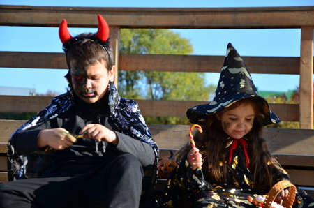 children in halloween costumes on holiday a girl in a witch suit a black hat on the head with black Halloween make-up, boy in devil halloween costume with horns zombie eating sweets jelly wormsの写真素材