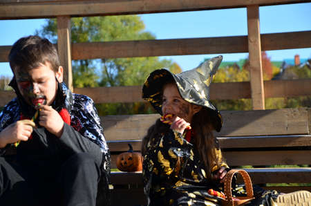 children in halloween costumes on holiday a girl in a witch suit a black hat on the head with black Halloween make-up, boy in devil halloween costume with horns zombie eating sweets jelly wormsの写真素材