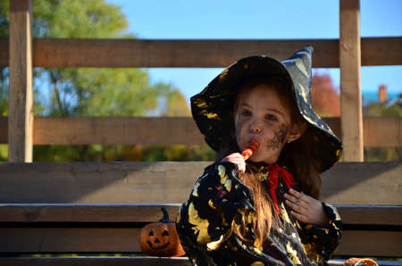 a girl in a witch suit and a black hat on the head with black Halloween make-up, zombie eating sweets jelly worms at a celebration of Halloweenの写真素材