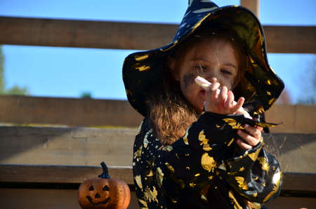 a girl in a witch suit and a black hat on the head with black Halloween make-up, zombie eating sweets jelly worms at a celebration of Halloweenの写真素材