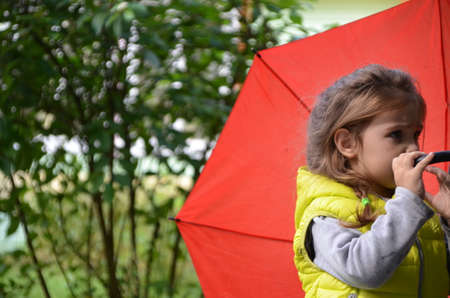 little funny cute girl walks in the rain with a red umbrella, in green rubber boots through the puddles. Laughs, has fun. girl wearing yellow waterproof coat playing on a warm autumn or summer dayの写真素材