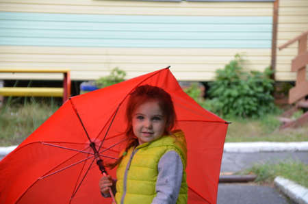 little funny cute girl walks in the rain with a red umbrella, in green rubber boots through the puddles. Laughs, has fun. girl wearing yellow waterproof coat playing on a warm autumn or summer dayの写真素材