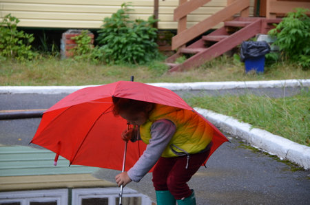 little funny cute girl walks in the rain with a red umbrella, in green rubber boots through the puddles. Laughs, has fun. girl wearing yellow waterproof coat playing on a warm autumn or summer dayの写真素材