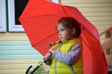 little funny cute girl walks in the rain with a red umbrella, in green rubber boots through the puddles. Laughs, has fun. girl wearing yellow waterproof coat playing on a warm autumn or summer dayの写真素材