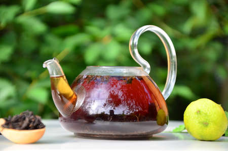 teapot full of tea on a background of green foliage on a wooden table, next to forest berries, raspberries and blueberries, lime or bergamot, mint. healthy summer drinkの写真素材