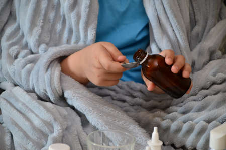 Sick child boy lying in bed with a fever, resting at home. a boy with a cold, treated. medicines, throat nose spray. sick kid with runny nose and fever heat lying on couch at home. drinking hot drinkの写真素材