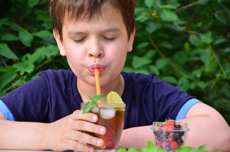 Child boy drinks natural lemonade summer drink, tea with wild berries, lime, raspberries and blueberries, summer vacation in park. refreshing natural drink lemonade Detox fruit infused water cocktailの写真素材