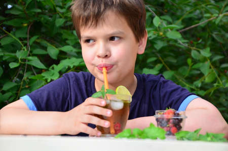 Child boy drinks natural lemonade summer drink, tea with wild berries, lime, raspberries and blueberries, summer vacation in park. refreshing natural drink lemonade Detox fruit infused water cocktailの写真素材