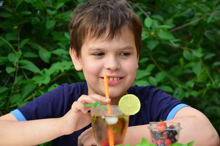 Child boy drinks natural lemonade summer drink, tea with wild berries, lime, raspberries and blueberries, summer vacation in park. refreshing natural drink lemonade Detox fruit infused water cocktailの写真素材