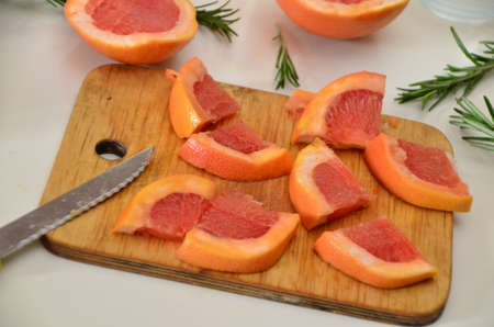 young lady in white dress is cutting grapefruit with big knife. rosemary on the table. to prepare detox water or shake.の写真素材