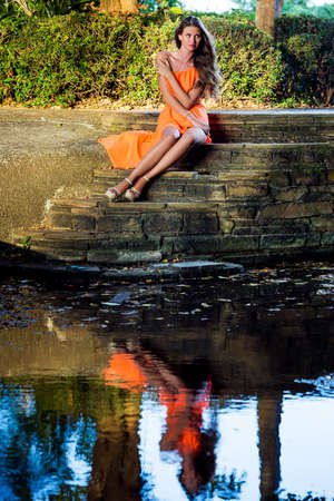 Young woman in orange dress sitting on stairs in the tropical park with reflection in waterの写真素材