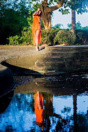 Young woman in orange dress posing in the tropical park with reflection in waterの写真素材