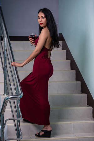 Beautiful asian woman in long evening dress with glass of red wine standing on a stepsの写真素材