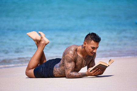 Athletic tattooed guy reading book on the sand at the tropical beachの写真素材