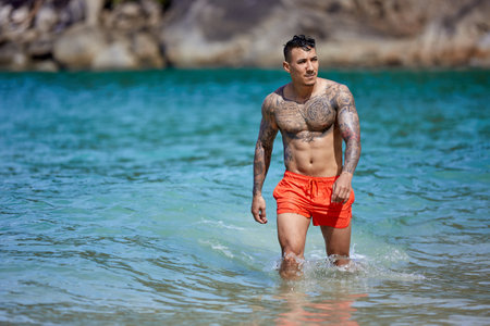 Athletic shirtless tattooed guy in red shorts walking through the water at the tropical beachの写真素材