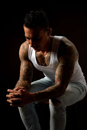 Portrait of athletic young tattooed guy in white undershirt and blue jeans. Black background. Studio shotの写真素材