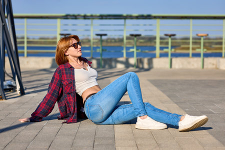 A fashionable redhead woman enjoying an autumn evening in the city. She's wearing a trendy red jacket, a white t-shirt, and denim jeans, making a bold fashion statementの写真素材