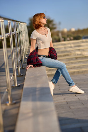 A fashionable redhead woman enjoying an autumn evening in the city. She's wearing a trendy red jacket, a white t-shirt, and denim jeans, making a bold fashion statementの写真素材