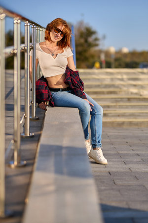 A fashionable redhead woman enjoying an autumn evening in the city. She's wearing a trendy red jacket, a white t-shirt, and denim jeans, making a bold fashion statementの写真素材