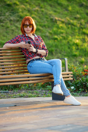 A fashionable redhead woman enjoying an autumn evening in the city. She's wearing a trendy red shirt and denim jeans, making a bold fashion statementの写真素材