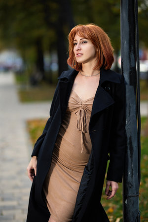 A fashionable redhead woman enjoying and smiling an autumn evening in the city. She's wearing a trendy black coat and beige dress, making a bold fashion statementの写真素材