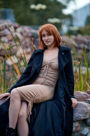 A fashionable redhead woman enjoying and smiling an autumn evening in the city. She's wearing a trendy black coat and beige dress, making a bold fashion statementの写真素材