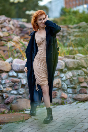 A fashionable redhead woman enjoying and smiling an autumn evening in the city. She's wearing a trendy black coat and beige dress, making a bold fashion statementの写真素材
