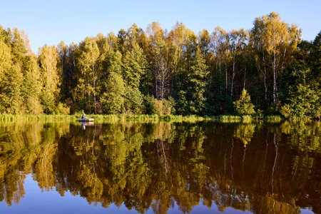 A lone fisherman in a boat on a peaceful lake, surrounded by dense green forest and reflected trees. Perfect for nature, outdoor activities, and fishing themesの写真素材